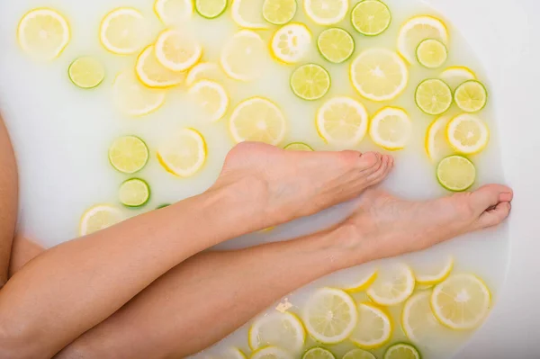 Unerkennbare Frau nimmt ein Milchbad mit Zitronen und Limetten. Zitrusbad. Körperpflege. Hautaufhellung. Weibliche Beine von oben. — Stockfoto