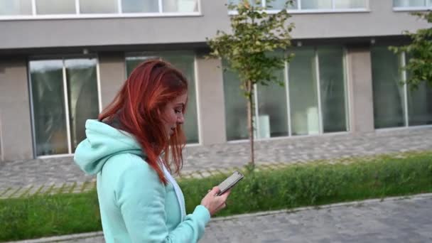 Młoda rudowłosa kobieta idzie ulicą i nagrywa filmik z selfie przez telefon. Piękna dziewczyna w bluzie ma wideo czat na smartfonie. — Wideo stockowe