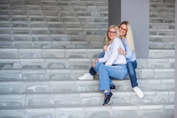 A beautiful blonde with glasses and her mature mother are sitting on the stairs outdoor. A well-groomed elderly woman hugs her adult daughter tightly. Love and family values. Mothers Day. — Stock Photo, Image
