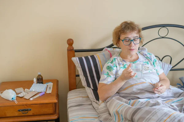 Sick old woman is lying at home in bed. A pensioner takes a cure for the disease and drinks a glass of water.