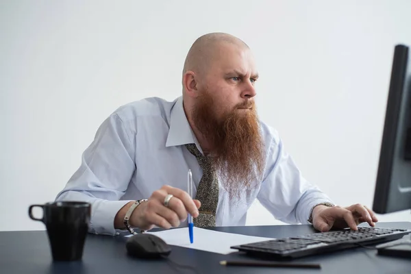 A bald man with a red beard in the office in a business suit works at the computer. The manager stares intently at the monitor, solves problems. — Stockfoto