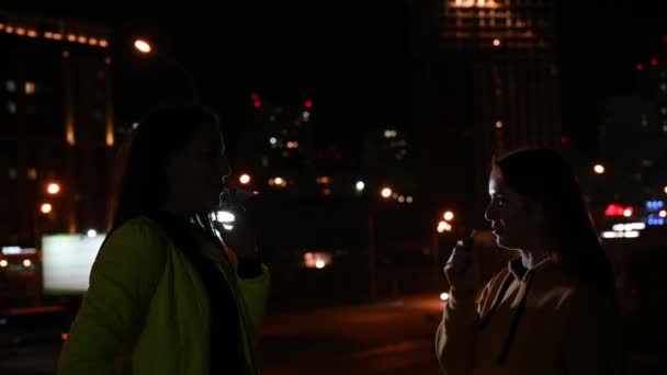 Twee vriendinnen roken op de achtergrond van de nachtstad. Vrouwen roken 's avonds laat sigaretten en vape. Slechte gewoonte. — Stockvideo