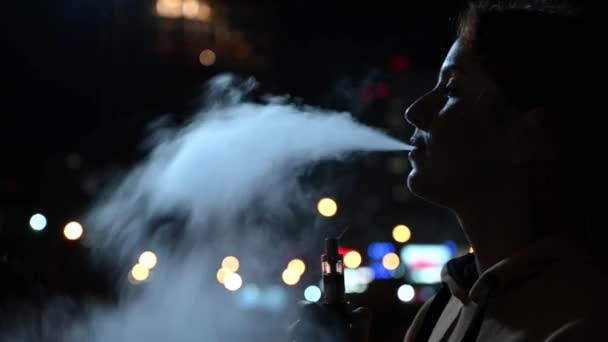 La chica fuma un cigarrillo electrónico contra el telón de fondo de la ciudad nocturna. Mujer disfrutando del vapor, vaporizador de vapor de nicotina. Mal hábito. — Vídeos de Stock