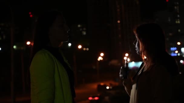 Två flickvänner röker i bakgrunden av nattstaden. Kvinnor röker cigaretter och ånga sent på kvällen. Dålig vana. — Stockvideo