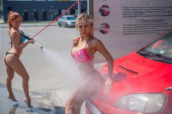 Chicas de lujo en bikinis seductores lavan un coche en un lavado de coches de la calle y el agua entre sí. Rubia y pelirroja con hermosas figuras lavan un coche rojo en un lavadero manual. . —  Fotos de Stock