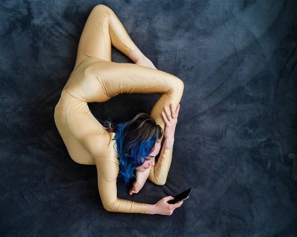 Samice akrobata leží na šedé pokrývce v zakřivené póze a používá mobil. Žena, která odpočívá na posteli v akrobatickém kousku a píše do telefonu.. — Stock fotografie