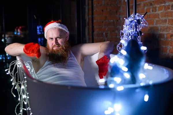 Vicces kép egy gonosz télapóról, aki karácsonyi fényekkel fürdik. Egy férfi vörös szakállal, télapó sapkában és alsóneműben egy koszorút tart a kezében.. — Stock Fotó