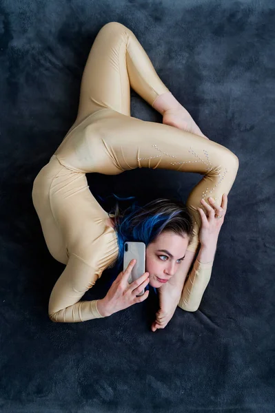 Eine weibliche Akrobatin liegt in geschwungener Pose auf einer grauen Bettdecke und benutzt ein Handy. eine Verrenkungskünstlerin, die sich bei einem akrobatischen Stunt auf einem Bett ausruht und per SMS telefoniert. — Stockfoto