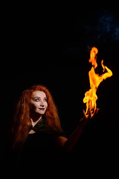 An ominous witch with long curly hair holds a magical fire. Tongues of flame on the palms of a red-haired smirking woman. Witchcraft.