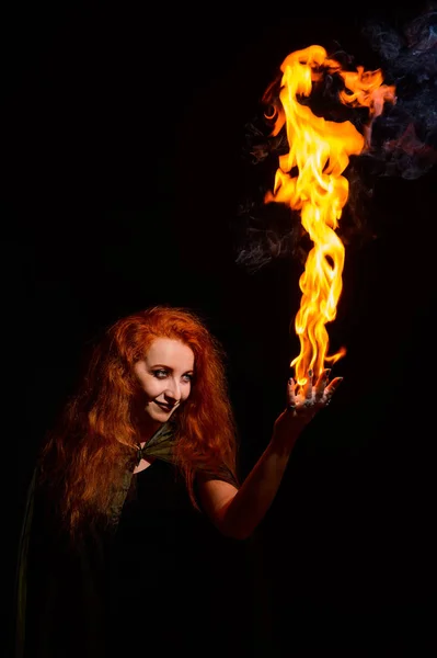 An ominous witch with long curly hair holds a magical fire. Tongues of flame on the palms of a red-haired smirking woman. Witchcraft.