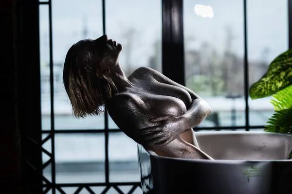Nackte Frau mit sehr großen Brüsten und silbernem Body im Badezimmer. Ein Mädchen mit einer extragroßen Oberweite mit Funkeln auf der Haut duscht. — Stockfoto
