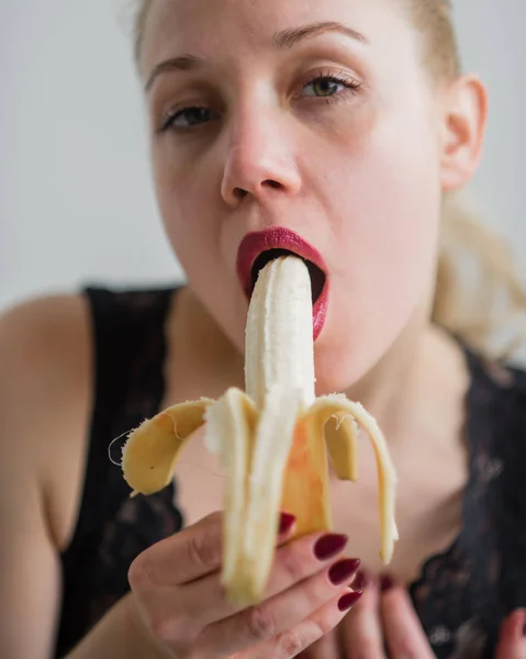 En blondin i svarta spets underkläder förföriskt suger och slickar en banan. Kvinna med sensuell röd imiterar oral njutning. — Stockfoto