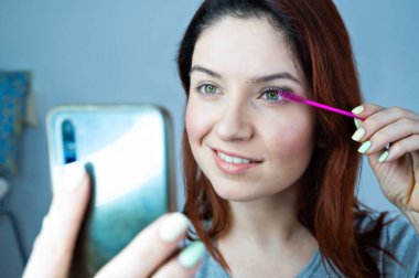 Güzel kızıl saçlı kadın kirpiklerini boyuyor ve selfie çekiyor. Kız maskara sürüyor ve ayna yerine telefona bakıyor..