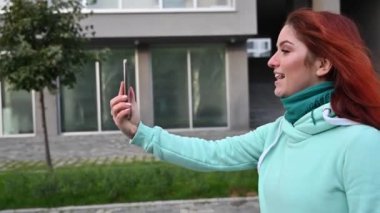 Genç kızıl saçlı bir kadın sokakta yürüyor ve telefonda bir selfie videosu kaydediyor. Sweatshirt 'lü güzel bir kız akıllı telefondan görüntülü konuşma yapar..