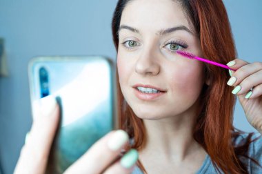 Güzel kızıl saçlı kadın kirpiklerini boyuyor ve selfie çekiyor. Kız maskara sürüyor ve ayna yerine telefona bakıyor..