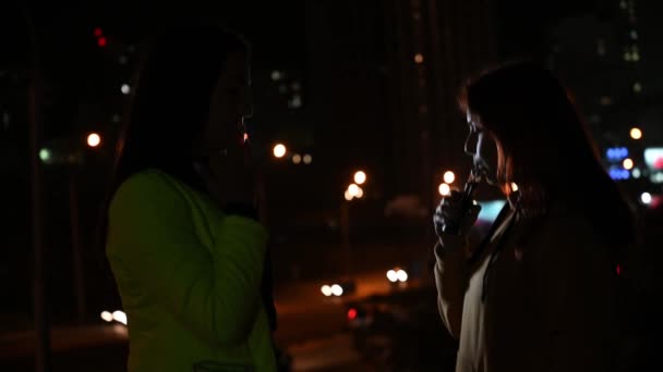 Dos novias fuman en el fondo de la ciudad nocturna. Las mujeres fuman cigarrillos y vapean tarde en la noche. Mal hábito. — Vídeos de Stock