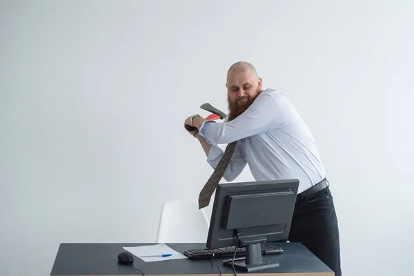 Злой лысый мужчина с рыжей бородой в офисе в деловом костюме разбивает топор компьютером. Менеджер с нервным срывом сломал монитор . — стоковое фото