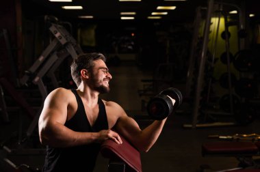 Gözlüklü, sakallı yakışıklı bir adam halterle pazılar için egzersiz yapıyor. Genç adam vücut geliştirmekle meşgul. Spor salonundaki antrenör kaslı kollarıyla gülümsüyor..