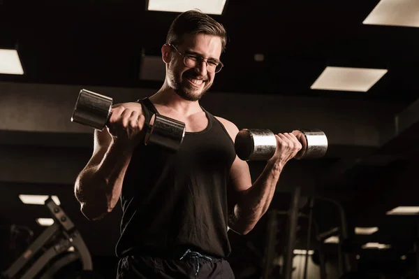 Egy férfi, akinek van erőfeszítése, bicepszeket és tricepszeket csinál súlyzóval. Dombornyomott testépítő mosolyog. Izmos fickó szemüveggel az edzőteremben. — Stock Fotó