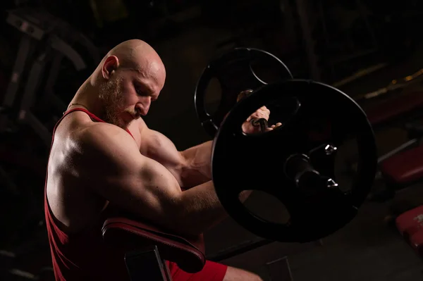Egy szakállas kopasz ember bicepszeket gyakorol egy súlyzóval. A fiatalember testépítéssel foglalkozik. Edző az edzőteremben izmos karokkal — Stock Fotó
