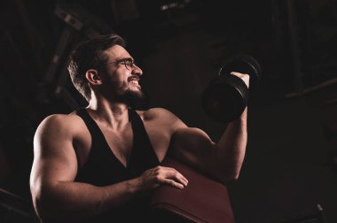 Gözlüklü, sakallı yakışıklı bir adam halterle pazılar için egzersiz yapıyor. Genç adam vücut geliştirmekle meşgul. Spor salonundaki antrenör kaslı kollarıyla gülümsüyor..