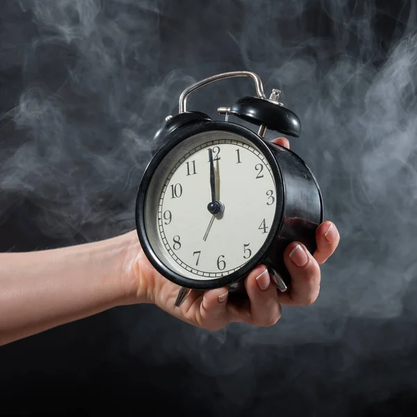 Close-up of a female hand holding a clock on a black background in smoke. Alarm clock at midnight in a mystical fog.