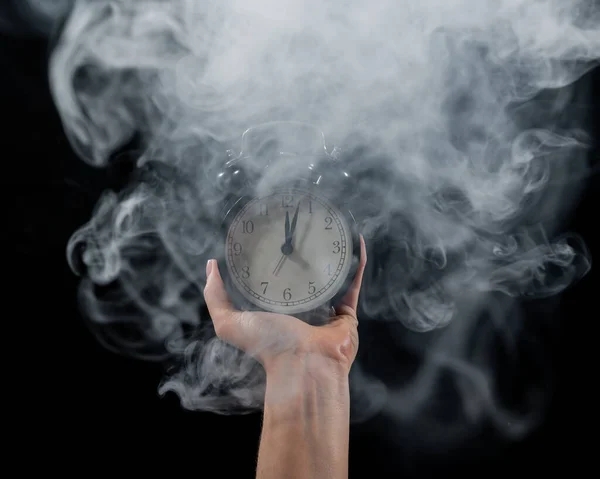 Primer plano de una mano femenina sosteniendo un reloj sobre un fondo negro en humo. Reloj despertador a medianoche en una niebla mística . —  Fotos de Stock