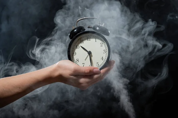 A woman holds an alarm clock in a studio full of smoke. White fog enveloped a round retro mechanical watch.