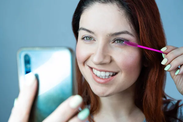 Mulher ruiva bonita pinta pestanas e toma uma selfie. A menina coloca rímel e olha para o telefone em vez de um espelho . — Fotografia de Stock