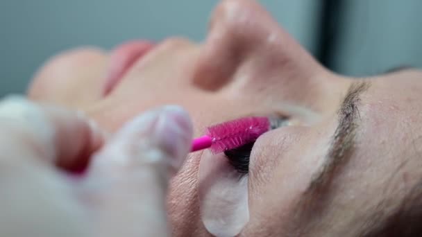 En kvinna ligger med slutna ögon i en skönhetsprocedur. Mästaren kammar förlängningsögonfransarna med en rosa borste. — Stockvideo