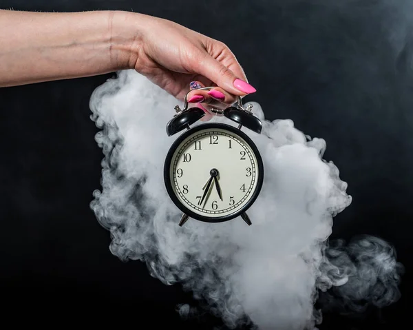 Una mujer sostiene un despertador en un estudio lleno de humo. Niebla blanca envolvió un reloj mecánico retro redondo . —  Fotos de Stock