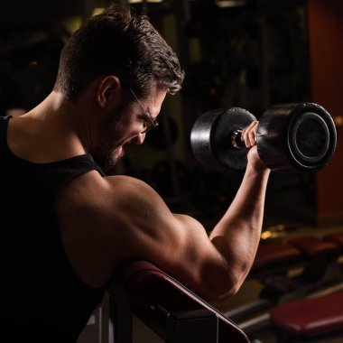 Gözlüklü yakışıklı bir adam halterle pazılar için egzersiz yapıyor. Adam vücut geliştirmekle meşgul. Spor salonunda kaslı kollarıyla antrenör..