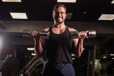 Çabalayan bir adam, pazılar ve trisepsler için halterli egzersiz yapar. Kabartmalı vücut geliştirici gülümsemesi. Spor salonunda gözlüklü kaslı adam..