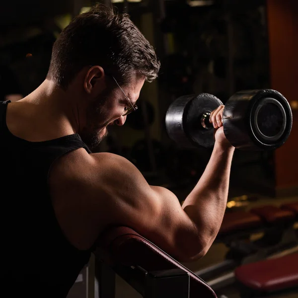 Egy jóképű, szemüveges férfi, aki bicepszre gyakorol egy súlyzóval. A fickó testépítéssel foglalkozik. Edző az edzőteremben izmos karokkal. — Stock Fotó