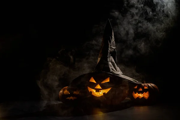 Drei gruselig grinsende Halloween-Kürbisse leuchten im Dunkeln im Nebel. Jack-o-Laterne mit Hexenhut auf schwarzem Hintergrund in Rauch. — Stockfoto