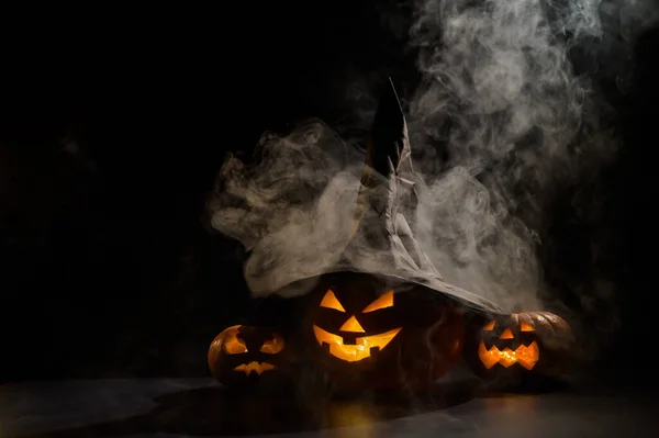 Trzy przerażające Halloween uśmiechające się dynie świecą w ciemności pośród mgły. Jack-o-latarnia w kapeluszu czarownicy na czarnym tle w dymie. — Zdjęcie stockowe