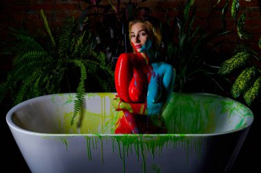 Beautiful woman with very big breasts takes a bath. Red and blue paint flows down the naked body of a girl in the bathroom. Creative body art. clipart