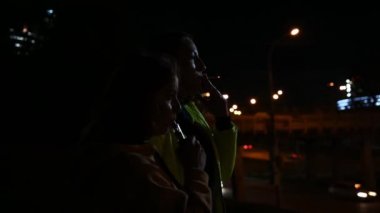 Gece şehrinin arka planında iki kız arkadaş sigara içiyor. Kadınlar gece geç saatlerde sigara ve sigara içerler. Kötü alışkanlık..