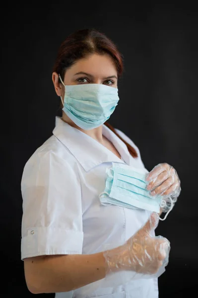 A young female doctor in uniform holds a medical mask on a black background. Nurse shows thumb ok. Coronovirus protection.