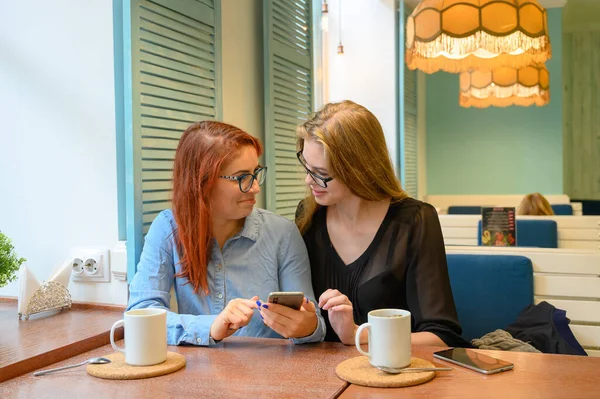 Two excited young girls using mobile phones, sitting in a cafe and pointing finger. Red-haired woman in glasses shows her friend funny photos on a smartphone girlfriend blonde. They drink coffee. — Stock Photo, Image