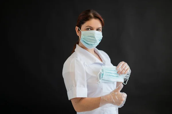 Una joven doctora de uniforme sostiene una máscara médica sobre un fondo negro. Enfermera muestra pulgar ok. Protección contra Coronovirus . —  Fotos de Stock