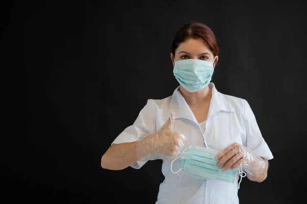 Una joven doctora de uniforme sostiene una máscara médica sobre un fondo negro. Enfermera muestra pulgar ok. Protección contra Coronovirus . —  Fotos de Stock