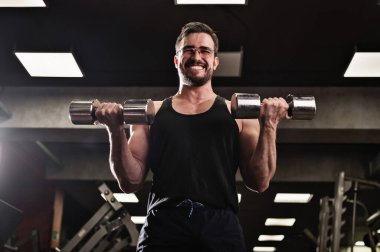 Çabalayan bir adam, pazılar ve trisepsler için halterli egzersiz yapar. Kabartmalı vücut geliştirici gülümsemesi. Spor salonunda gözlüklü kaslı adam..