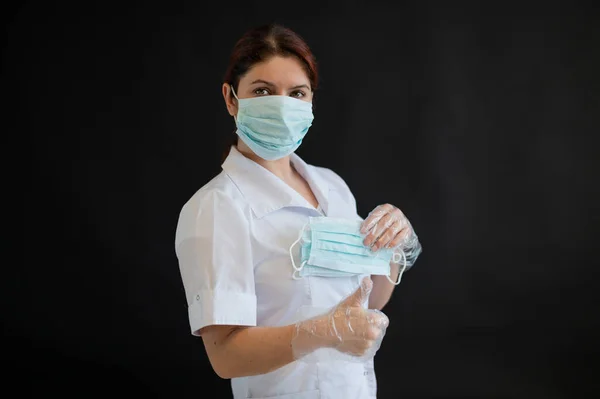 Una joven doctora de uniforme sostiene una máscara médica sobre un fondo negro. Enfermera muestra pulgar ok. Protección contra Coronovirus . —  Fotos de Stock