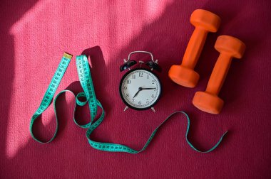 Pembe yoga minderinde iki turuncu tünel mezurası ve çalar saat. Spor yapma ve yazın kilo verme zamanı geldi. Evde ekipmanlı spor.