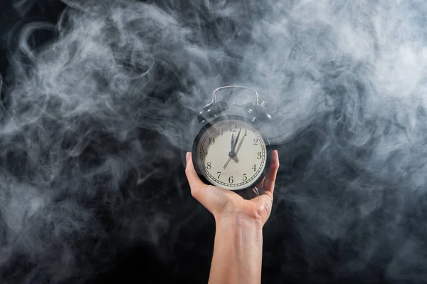 Close-up of a female hand holding a clock on a black background in smoke. Alarm clock at midnight in a mystical fog.