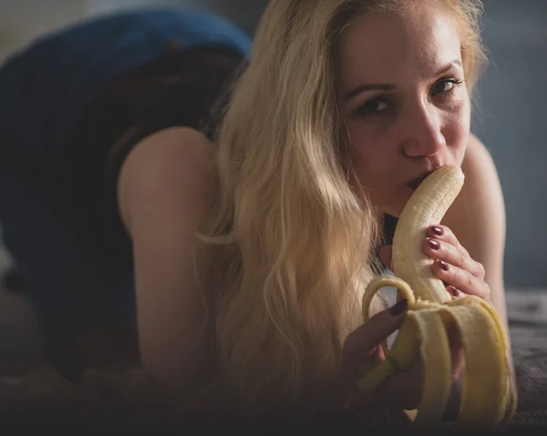 Eine Blondine in schwarzen Spitzenunterwäsche saugt und leckt verführerisch eine Banane. Frau mit sinnlichem Rot imitiert orales Vergnügen. — Stockfoto