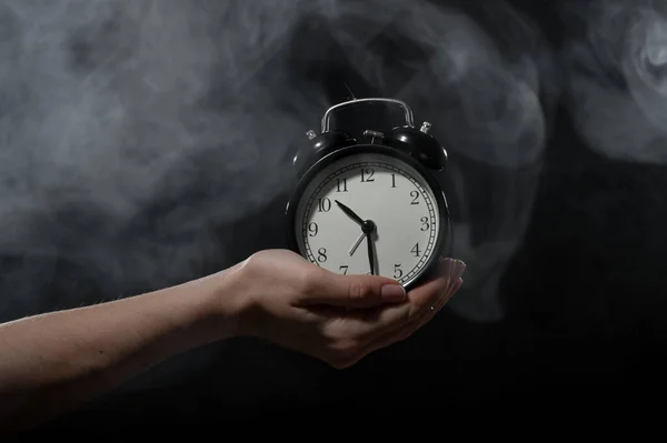 Una mujer sostiene un despertador en un estudio lleno de humo. Niebla blanca envolvió un reloj mecánico retro redondo . —  Fotos de Stock