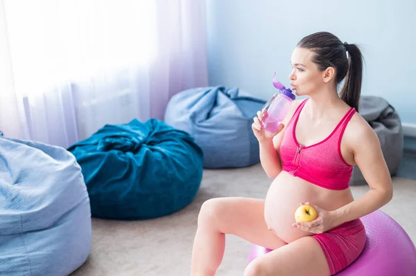 Zwangere vrouw zonder gezicht die een appel eet na yoga. Verwachte moeder bijt fruit en drinkt water na Pilates. Meisje zittend op een fitness bal. — Stockfoto