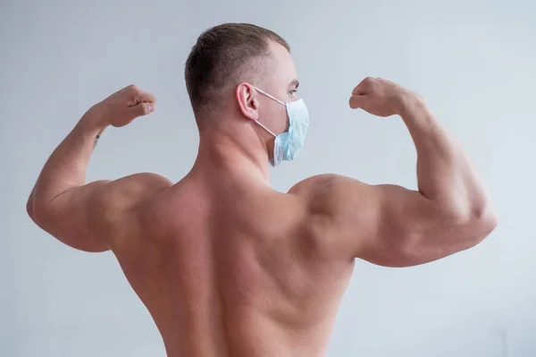 Homme bodybuilder dans un masque médical se tient avec son dos. Il va faire du sport en quarantaine. Protection respiratoire. Montre biceps . — Photo